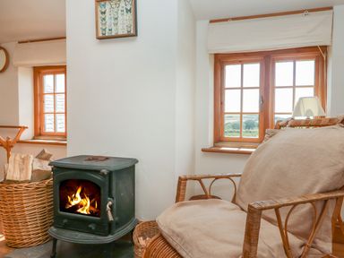 A room with a wood burning stove a wicker chair a basket of firewood and two windows at Higher Roskorwell Cottage in St Keverne