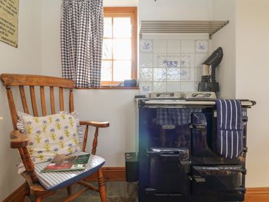 A wooden chair with cushions and a magazine next to a stove with tiled backsplash and a small window with checkered curtains at Higher Roskorwell Cottage in St Keverne