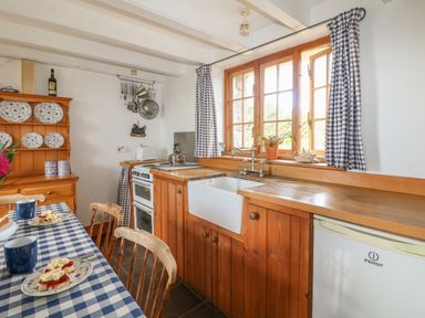 A kitchen with a checkered tablecloth on a table wooden chairs a farmhouse sink wooden cabinets and utensils hanging above a stove at Higher Roskorwell Cottage in St Keverne