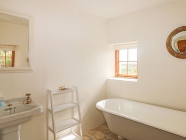 A bathroom with a freestanding bathtub a window a sink with a mirror and a small white shelf unit at Higher Roskorwell Cottage in St Keverne