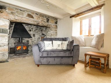 A living room with a stone fireplace and wood burning stove a gray sofa with cushions a window seat with pillows and a wooden side table at Higher Roskorwell Cottage in St Keverne