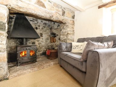 A room with a stone fireplace a burning wood stove and a gray sofa with cushions at Higher Roskorwell Cottage in St Keverne
