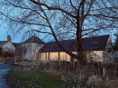 A stone building with lit windows and a tree in front at dusk at High Cogges Farm Holiday Cottages - The Cart Shed High Cogges near Witney