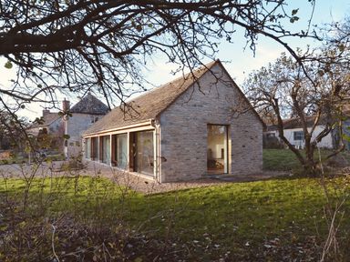 Stone building with large windows and a pitched roof in a garden with grass and trees at High Cogges Farm Holiday Cottages - The Cart Shed in High Cogges near Witney