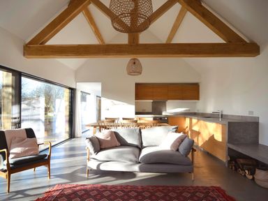 An open plan living room and kitchen with wooden beams a grey sofa a chair a dining table and large windows at High Cogges Farm Holiday Cottages - The Cart Shed in High Cogges near Witney