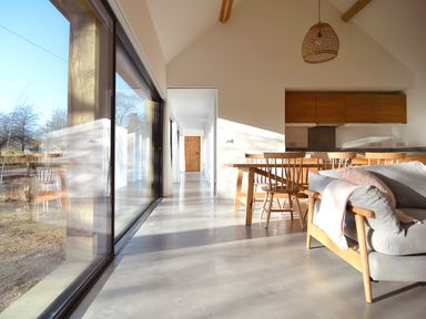 A dining area with wooden chairs and table next to a grey sofa in a room with a large glass window at High Cogges Farm Holiday Cottages - The Cart Shed in High Cogges near Witney