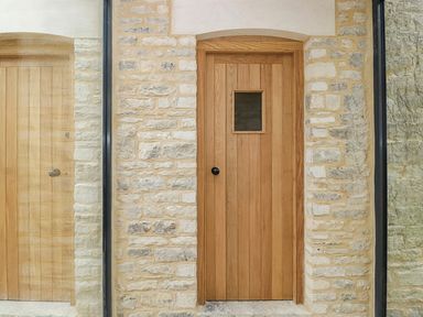 A wooden door with a small square window in a stone wall and a glass panel reflecting the door at High Cogges Farm Holiday Cottages - The Cart Shed in High Cogges near Witney