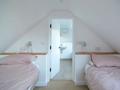 A bedroom with two beds facing each other with pink bedding a door partially open showing a bathroom with a sink at High Cogges Farm Holiday Cottages - The Cart Shed High Cogges near Witney