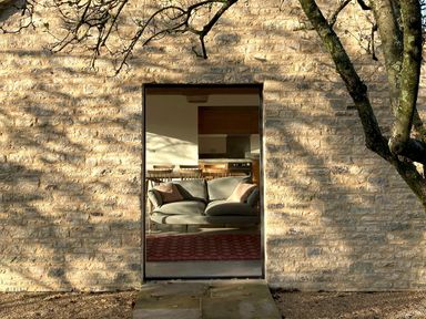 A stone wall with an open doorway showing a green sofa with cushions and wooden chairs around a table inside a room at High Cogges Farm Holiday Cottages - The Cart Shed in High Cogges near Witney