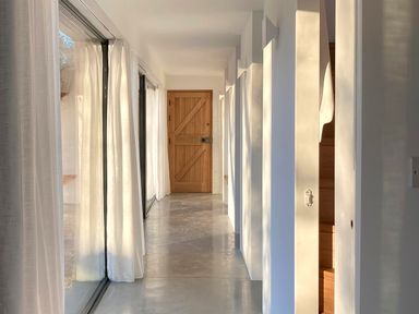 A hallway with concrete floor glass sliding doors and white curtains leading to a wooden door at High Cogges Farm Holiday Cottages - The Cart Shed High Cogges near Witney