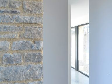A stone wall next to a white wall with a doorway leading to a room with large windows