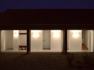 A building exterior at night with three illuminated rooms showing white walls wooden bench baskets and stairs at High Cogges Farm Holiday Cottages - The Cart Shed High Cogges near Witney