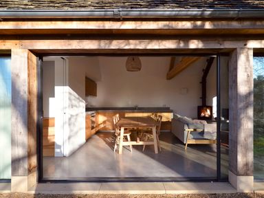 A kitchen and living area with a wooden dining table and chairs a gray sofa and a wood burning stove at High Cogges Farm Holiday Cottages - The Cart Shed in High Cogges near Witney
