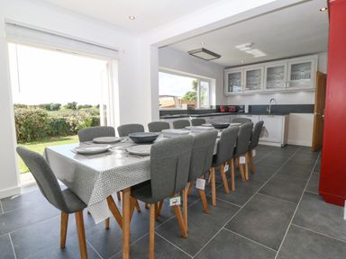 A dining area with a long table set with plates and bowls and multiple grey chairs in a kitchen with white cabinets at Glanffraw Aberffraw Ty Croes