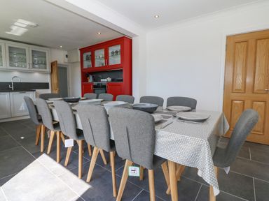 A dining area with a long table covered by a tablecloth and set with plates and bowls surrounded by gray chairs in a kitchen with white and red cabinets at Glanffraw in Aberffraw Ty Croes