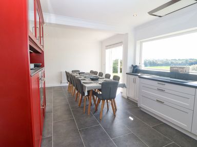 A dining area with a long table set with dishes and grey chairs next to red and white kitchen cabinets at Glanffraw in Aberffraw Ty Croes