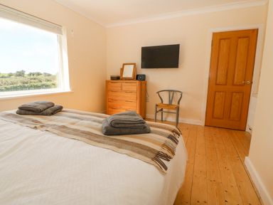 A bedroom with a bed folded towels on it a wooden dresser a chair a wall mounted tv and a wooden door at Glanffraw in Aberffraw Ty Croes