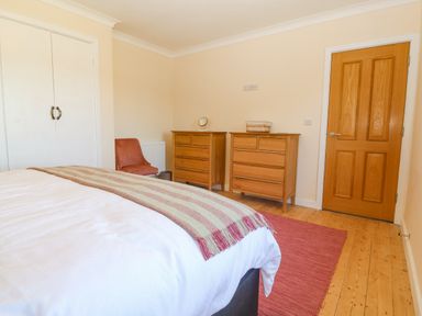 A bedroom with a bed a striped blanket two wooden dressers a red chair a wooden door and a closet at Glanffraw Aberffraw Ty Croes