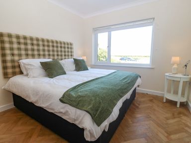 A bedroom with a double bed green cushions and headboard white side table with lamp and window showing outdoor view at Glanffraw Aberffraw Ty Croes