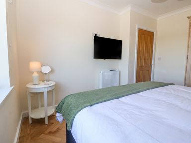 A bedroom with a bed covered by a white duvet and green throw a small round table with a lamp and mirror a wall mounted television a wooden door and a radiator at Glanffraw Aberffraw Ty Croes
