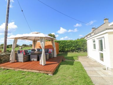 A backyard with a wooden deck covered by a canopy with outdoor seating and cushions at Glanffraw in Aberffraw Ty Croes