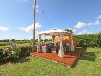 An outdoor wooden deck with a gazebo covering a seating area with cushions and a table surrounded by grass at Glanffraw in Aberffraw Ty Croes