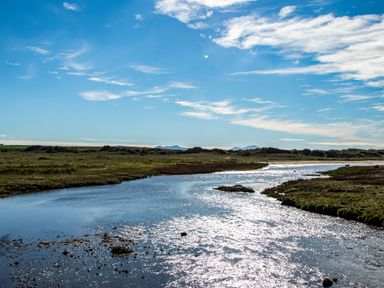 A river flowing through a grassy flatland under a blue sky with scattered clouds at Glanffraw Aberffraw Ty Croes