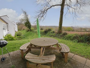A wooden round picnic table with benches on a patio near a tree and a grassy area at Hembry Meadows in Honicombe Park near Gunnislake