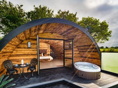 A wooden pod with an open door showing a bed inside a patio with a small table and chairs and a hot tub at The Piglet Pod in Llanarth