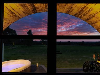 A view through a window showing a lit hot tub on a deck and a grassy field with trees at sunset at The Piglet Pod in Llanarth