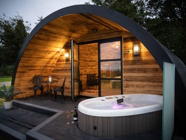 A wooden curved cabin with a hot tub two chairs and a small table at The Piglet Pod in Llanarth