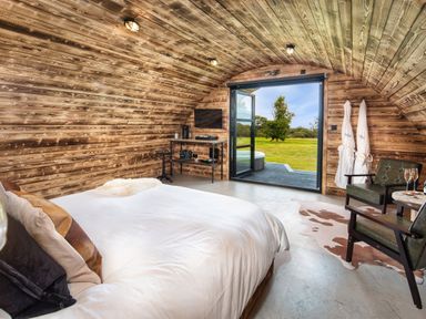 A bedroom with a large bed a small table two chairs robes hanging on the wall and a door open to a grassy outdoor area at The Piglet Pod in Llanarth