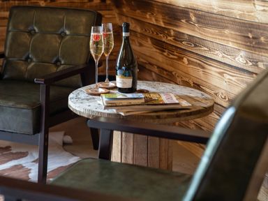 A wooden round table with a bottle of prosecco and two glasses with raspberries next to two green leather chairs at The Piglet Pod in Llanarth