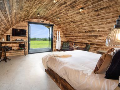A bedroom with wooden walls a bed two chairs a television and open doors showing a green field at The Piglet Pod in Llanarth