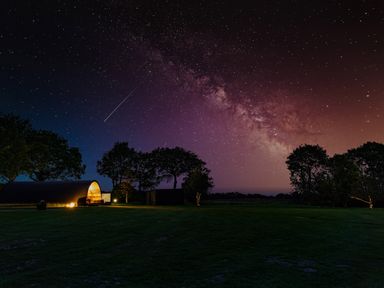 A night sky filled with stars and the Milky Way over a field with trees and a lit dome structure at The Piglet Pod in Llanarth