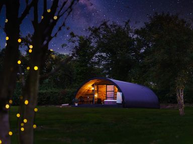 A lit curved cabin with outdoor seating and a hot tub in a grassy area with trees and starry sky at The Piglet Pod in Llanarth