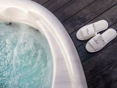 A bubbling white jacuzzi tub next to a pair of white slippers on a dark wooden floor at The Piglet Pod in Llanarth