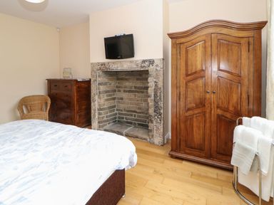 A bedroom with a bed a wicker chair a wooden chest of drawers a stone fireplace with a mounted TV a wooden wardrobe and towels on a rack at Houghton North Farm Cottage in Heddon-on-the-Wall near Hexham
