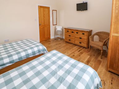 A bedroom with two beds covered with blue and white checkered bedspreads a wooden chest of drawers a wicker chair a small wall-mounted television a wooden door and a towel rack at Houghton North Farm Cottage in Heddon-on-the-Wall near Hexham