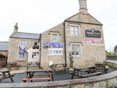 An outdoor seating area with wooden picnic tables in front of a stone building with signs reading Three Tuns and Howay The Lads at Houghton North Farm Cottage in Heddon-on-the-Wall near Hexham