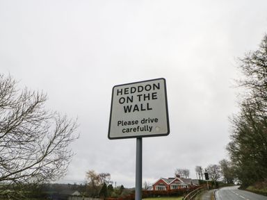 A road sign that says Heddon on the Wall Please drive carefully next to a road with trees and houses visible