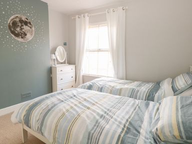 A bedroom with two single beds a chest of drawers with a mirror a window with white curtains and a moon and stars wall decal at Gull's Nest in Pakefield