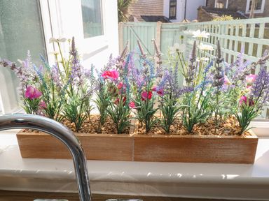 A kitchen window with two wooden planters of artificial flowers and a metal faucet at Gull's Nest in Pakefield
