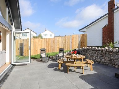 A patio with a wooden picnic table and two black chairs near a stone wall and wooden fence at The Herun in Rhosneigr