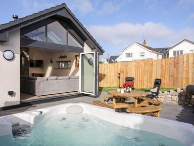 An outdoor patio area with a hot tub wooden picnic table and chairs next to a living room with gray sofa at The Herun in Rhosneigr