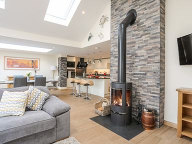 An open living room and kitchen area with a gray sofa two patterned pillows a wood burning stove with a stone chimney and a kitchen island with bar stools at The Herun in Rhosneigr
