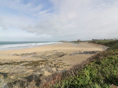 A sandy beach with rocks and some people by the ocean under a partly cloudy sky with green vegetation in the foreground at Newquay Fistral Beach View in Newquay