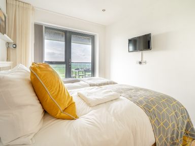 A bedroom with a double bed yellow pillows mounted television and glass doors leading to a balcony at Newquay Fistral Beach View in Newquay