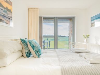 A bedroom with a bed, two blue pillows, folded towels, a chair, and a table near glass doors showing an outdoor view at Newquay Fistral Beach View in Newquay