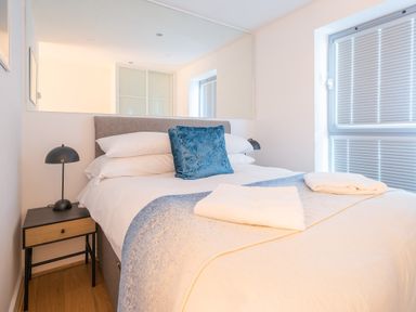 A bedroom with a bed a blue pillow a bedside table with a lamp and a window with blinds at Newquay Fistral Beach View in Newquay